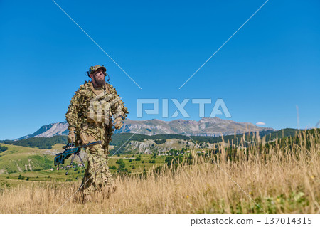 Soldier Sniper In Camouflage Ghillie Suit Patrolling Mountain Meadow Under Clear Blue Sky 137014315