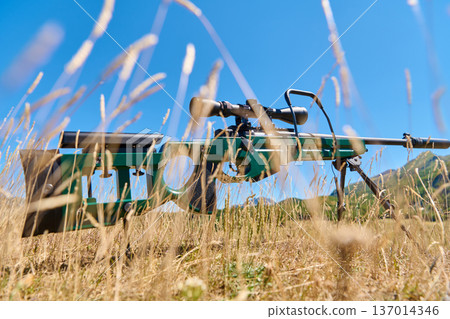 Sniper Rifle With Scope On Bipod Lying In Dry Grass In Open Mountain Field 137014346