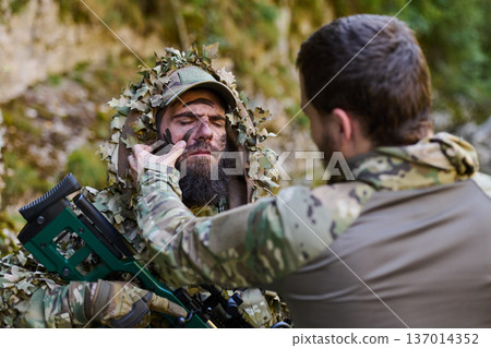 Soldier Applying Face Paint to Ghillie-Clad Sniper During Camouflage Preparation in Woodland Soldier Applying Face Paint to Ghillie-Clad Sniper During Camouflage Preparation in Woodland 137014352