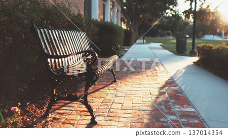 Vintage-Filtered Close-Up of an Empty Black Cast Iron Park Bench Resting on a Brick Sidewalk as the Warm Sunset Light Casts Long Shadows 137014354