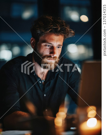 Intense Close-Up Portrait of a Focused, Bearded Man Working Late at Night on His Laptop in a Dimly Lit Modern Office or Creative Workspace 137014355