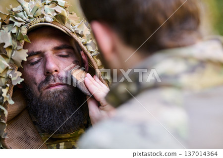 Soldier Applying Camouflage Face Paint While Preparing Ghillie Hood and Tactical Gear in Woodland 137014364