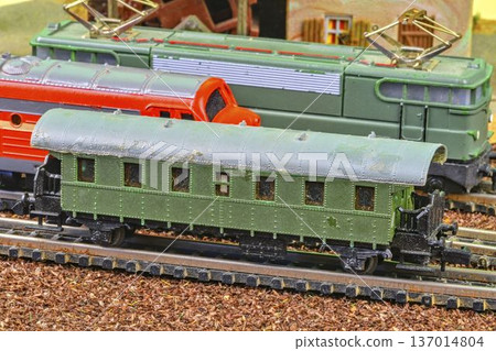 Vintage model train cars lined up on tracks, detailed miniature railway scene. Close-up view of weathered, green, and red model train engines and passenger cars on a miniature railway track with Vintage model train cars lined up on tracks, detailed miniature railway scene. Close-up view of weathered, green, and red model train engines and passenger cars on a miniature railway track with 137014804