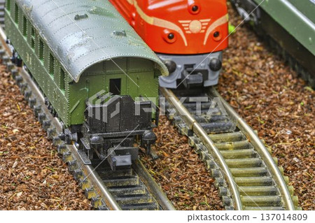 Close-up of two miniature train cars on a model railway track with wooden ballast. Detailed view of a green passenger car and a red locomotive on a curved track, showcasing intricate model railway Close-up of two miniature train cars on a model railway track with wooden ballast. Detailed view of a green passenger car and a red locomotive on a curved track, showcasing intricate model railway 137014809