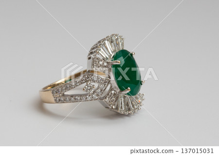 A gold ring featuring an oval cut green emerald surrounded by baguette cut diamonds on a clean white background. A gold ring featuring an oval cut green emerald surrounded by baguette cut diamonds on a clean white background. 137015031