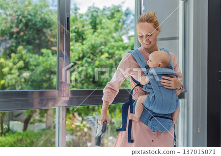 Woman vacuuming a room while carrying her baby in an ergonomic carrier. Modern motherhood, multitasking parent, home chores with infant, balance of childcare and housework concept. 137015071