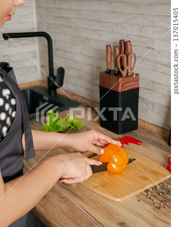 Girl cuts red pepper on a wooden board. Location of the kitchen. Girl cuts red pepper on a wooden board. Location of the kitchen. 137015405