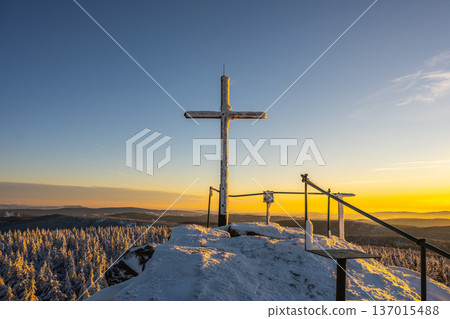 A cross stands at the summit rock formation of Jizera Mountain. Snow covers the ground and trees, and the sun sets in the background, creating a scenic winter view. 137015488