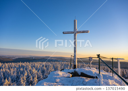 A cross stands atop the summit rock formation of Jizera Mountain. Snow covers the ground and trees, shining under the winter sunlight. The view stretches over distant hills and forests. A cross stands atop the summit rock formation of Jizera Mountain. Snow covers the ground and trees, shining under the winter sunlight. The view stretches over distant hills and forests. 137015490