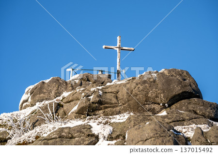 A wooden cross stands atop the rocky summit of Jizera Mountain. Snow covers the rocks under a bright blue sky in winter. This spot offers a clear view of the landscape below. A wooden cross stands atop the rocky summit of Jizera Mountain. Snow covers the rocks under a bright blue sky in winter. This spot offers a clear view of the landscape below. 137015492