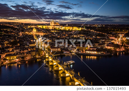 An aerial view of Prague at night. The Vltava River flows beneath the Charles Bridge while the castle is illuminated in the distance. Buildings and streets shine with lights. 137015493