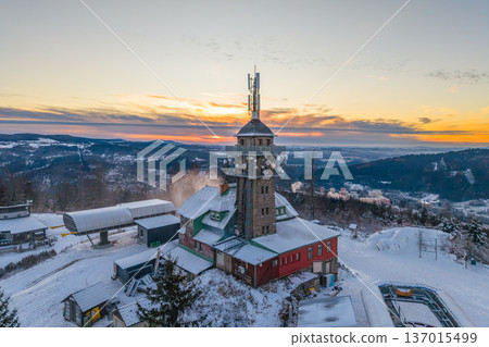 Lookout tower stands on Tanvaldsky Spicak mountain in winter. Snow covers structures as sunset lights the sky. Skis and snowboards are visible in the area. Lookout tower stands on Tanvaldsky Spicak mountain in winter. Snow covers structures as sunset lights the sky. Skis and snowboards are visible in the area. 137015499