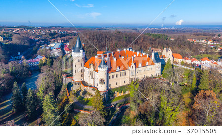 Aerial view shows Zleby Castle surrounded by trees and buildings. The castle features distinct towers and orange roofs. Green areas and clear sky are visible on a bright day. Aerial view shows Zleby Castle surrounded by trees and buildings. The castle features distinct towers and orange roofs. Green areas and clear sky are visible on a bright day. 137015505