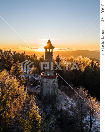 The lookout tower in the Jizera Mountains stands tall as the sun sets. Trees surround the tower, and the winter landscape glows with golden light as day turns to night. 137015507