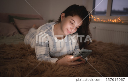 Young girl lies on a bed and looks at her phone during the evening, surfing on internet before sleep 137016067