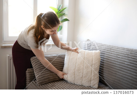 Young girl adjusts pillows on a couch near a window on a bright day, making the couch look neat 137016073