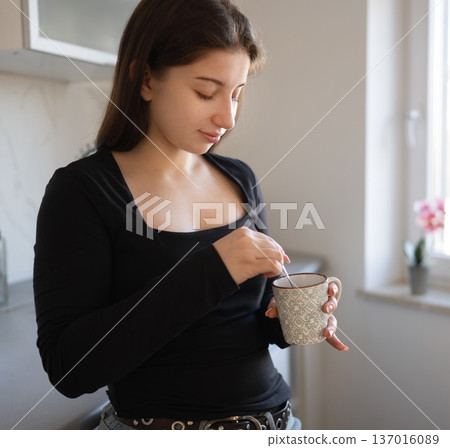 Girl stirs a drink in a cup while standing at a kitchen counter. Natural light comes in through the window nearby. 137016089