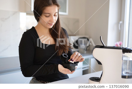 Young girl preparing to brew coffee with coffee capsules in kitchen during morning time. Young girl preparing to brew coffee with coffee capsules in kitchen during morning time. 137016090