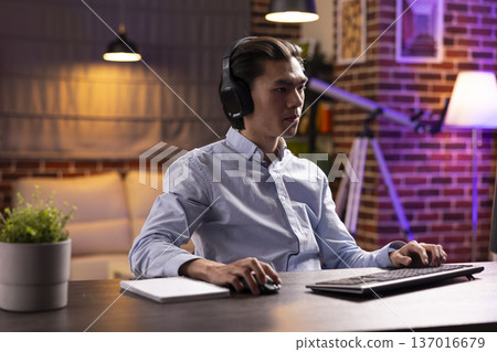 Young male professional listening to music while working remotely on computer. Businessman with headphones, enjoying balanced lifestyle, reading emails and finishing tasks in apartment home office. 137016679