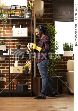 Smiling african american housewife stands near shelves, cleaning vase with cloth while listening to music. Black woman wearing headphones and gloves, enjoys doing household chores in brick wall home. 137016683