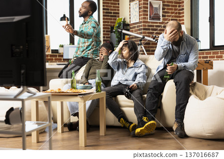 Frustrated friends sit with head in hand after losing game, showing disappointment and annoyance. Group of young adults in casual apartment reacts with sadness after tournament defeat. Frustrated friends sit with head in hand after losing game, showing disappointment and annoyance. Group of young adults in casual apartment reacts with sadness after tournament defeat. 137016687