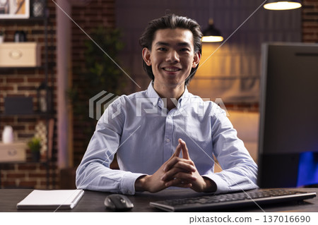 Smiling entrepreneur at home desk, excited to complete remote work. Handsome asian man sits with hands clasped, looking at camera in home office, ready to focus on evening online projects. Smiling entrepreneur at home desk, excited to complete remote work. Handsome asian man sits with hands clasped, looking at camera in home office, ready to focus on evening online projects. 137016690