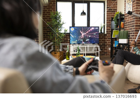Young female gamer enjoys gaming session on television at home with friends. Diverse people sitting comfortably in living room, playing video games on tv screen with wireless controllers. 137016693