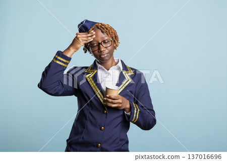 Tired flight attendant rubs forehead and holds warm cup of coffee, trying to regain energy after busy schedule. Weary air hostess lifts drink and massages head gently, reflecting exhaustion from work. Tired flight attendant rubs forehead and holds warm cup of coffee, trying to regain energy after busy schedule. Weary air hostess lifts drink and massages head gently, reflecting exhaustion from work. 137016696
