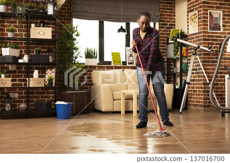 Housewife mopping in modern apartment, wearing casual clothes and cleaning hardwood floors in cozy living room on sunny afternoon. Black woman maintaining hygiene at home with mop and bucket. Housewife mopping in modern apartment, wearing casual clothes and cleaning hardwood floors in cozy living room on sunny afternoon. Black woman maintaining hygiene at home with mop and bucket. 137016700