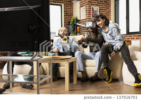 Focused black woman plays 3D game tournament with VR headset as cheerful white female friend supports. Diverse group enjoying weekend hangout in cozy apartment, cheering and watching gameplay. Focused black woman plays 3D game tournament with VR headset as cheerful white female friend supports. Diverse group enjoying weekend hangout in cozy apartment, cheering and watching gameplay. 137016701