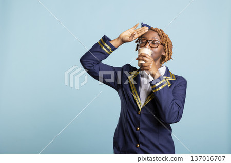 Stressed airline stewardess drinks from takeaway cup while gently touching forehead, expressing exhaustion and relief. Black female cabin crew member having cup of coffee during short break. 137016707