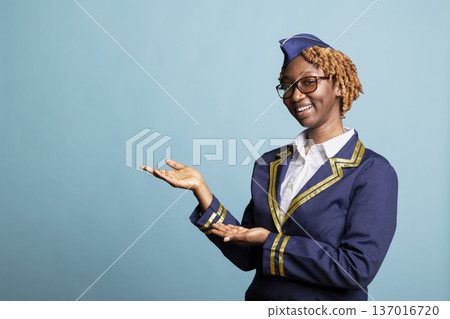 Cheerful air hostess stands against blue background, gesturing toward blank mockup space. Female flight attendant promotes airline offers, travel deals and professional aviation services in studio. Cheerful air hostess stands against blue background, gesturing toward blank mockup space. Female flight attendant promotes airline offers, travel deals and professional aviation services in studio. 137016720
