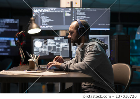 Caucasian software developer works intently on laptop, wearing headphones as monitors glow with technical data. Freelance programmer codes in tech office, reflecting concentration and digital workflow 137016726