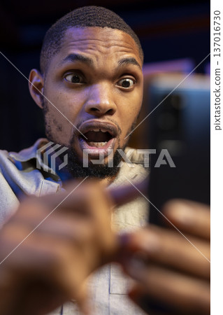 Shocked african american person reading surprising news on his smartphone, sitting in his living room with mouth open. Young adult being surprised while he holds mobile phone. Close up. Shocked african american person reading surprising news on his smartphone, sitting in his living room with mouth open. Young adult being surprised while he holds mobile phone. Close up. 137016730