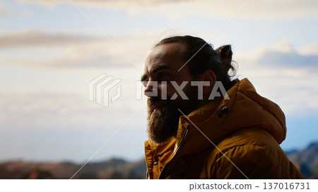 Victorious hiker relaxing at the mountain summit after climbing the top, proud of personal accomplishment. Young man sitting on the rocks and enjoying the peaceful surroundings. Camera B. 137016731