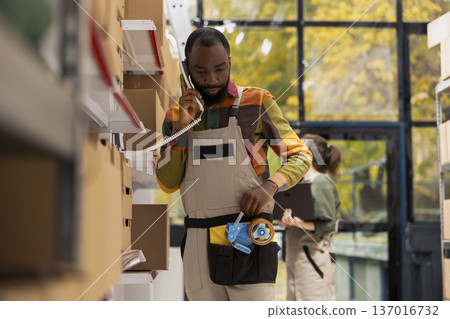 Black employee handling a landline phone call to check inventory lists and shipment details, preparing parcels and managing in-house logistics for online retail. E-business distribution. 137016732