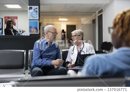 Senior physician reviews digital medical results with male patient, calmly discussing recovery plans and offering guidance. Female medic holds laptop and sits beside older man in clinic lobby. 137016771