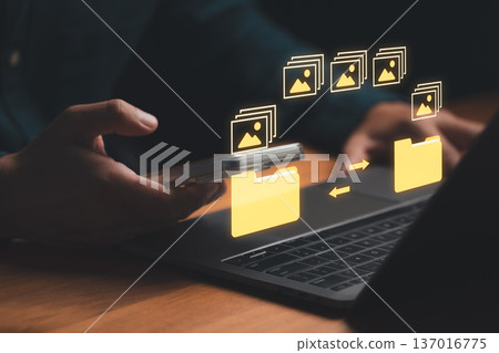 Man holding mobile phone with digital image file icon and a folder graphic between the phone and a laptop, representing photo transfer, cloud backup, data syncing, and cross device file management. 137016775