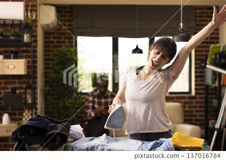 White woman ironing clothes and singing in brick wall apartment. Joyful female multitasks, dancing with one hand in air while removing wrinkles from pile of shirts during housework. White woman ironing clothes and singing in brick wall apartment. Joyful female multitasks, dancing with one hand in air while removing wrinkles from pile of shirts during housework. 137016784