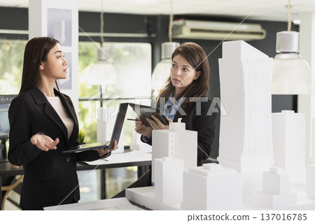 Diverse female designers with tablet and laptop, comparing between digital designs and 3d printed building layout. Professional women architects ensure accurate measurements of construction project. 137016785