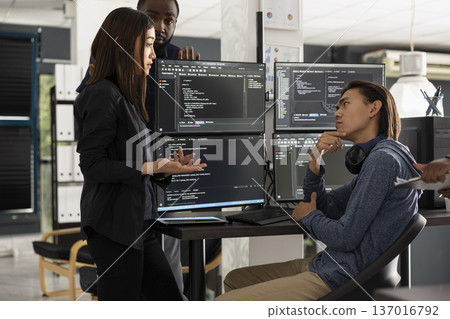 Male and female software developers stand by computer monitors while brainstorming coding project for startup. Diverse team collaborate on refining HTML scripts and building modern website together. Male and female software developers stand by computer monitors while brainstorming coding project for startup. Diverse team collaborate on refining HTML scripts and building modern website together. 137016792