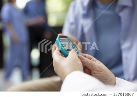 Close up of physician using digital glucometer to measure blood sugar levels in hospital lobby. Professional doctor uses glucose monitor and test strip for routine blood sugar testing on patient. Close up of physician using digital glucometer to measure blood sugar levels in hospital lobby. Professional doctor uses glucose monitor and test strip for routine blood sugar testing on patient. 137016807
