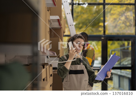 Woman worker in coveralls takes a phone call reviewing order lists and handling processing tasks for shipment service, storage facility operations and e-commerce express delivery. Woman worker in coveralls takes a phone call reviewing order lists and handling processing tasks for shipment service, storage facility operations and e-commerce express delivery. 137016810