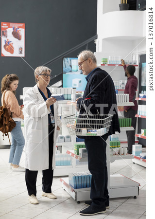 Woman pharmacist stands beside elderly male customer, explaining healthcare options near store shelves. The interaction highlights trust, expertise and personalized service in clean, modern pharmacy. Woman pharmacist stands beside elderly male customer, explaining healthcare options near store shelves. The interaction highlights trust, expertise and personalized service in clean, modern pharmacy. 137016814