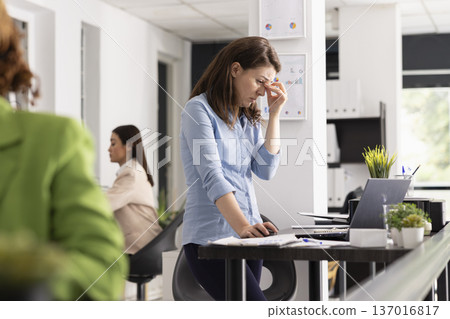 Stressed businesswoman touching face in frustration while reviewing documents. Professional female employee experiences pressure and fatigue during demanding workday in modern corporate office. Stressed businesswoman touching face in frustration while reviewing documents. Professional female employee experiences pressure and fatigue during demanding workday in modern corporate office. 137016817