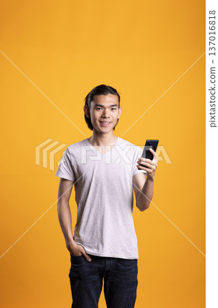 Happy male model in casual outfit poses with one hand in pocket, holding smartphone and looking at camera in modern studio. Young man texting friends on mobile device and browsing social media. Happy male model in casual outfit poses with one hand in pocket, holding smartphone and looking at camera in modern studio. Young man texting friends on mobile device and browsing social media. 137016818
