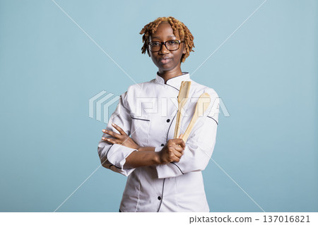 Focused female chef stands with arms crossed while holding wooden cooking utensils in studio. Young culinary professional expressing confidence, skill and discipline in food industry. 137016821