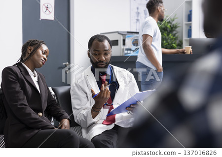 Male doctor reviews important medical paperwork with female visitor seated beside him in clinic waiting area. They discuss treatment details as other patients move around in busy healthcare lobby. Male doctor reviews important medical paperwork with female visitor seated beside him in clinic waiting area. They discuss treatment details as other patients move around in busy healthcare lobby. 137016826