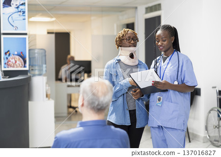 Friendly nurse with clipboard speaks to woman wearing neck brace in hospital lobby. Injured patient listens as healthcare worker explains treatment steps and answers questions in clinic waiting area. Friendly nurse with clipboard speaks to woman wearing neck brace in hospital lobby. Injured patient listens as healthcare worker explains treatment steps and answers questions in clinic waiting area. 137016827