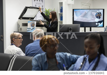Asian administrator in hospital lobby scans and prints medical documents. Female healthcare worker operates photocopier and printer, assisting doctors with patient records and administrative paperwork 137016841